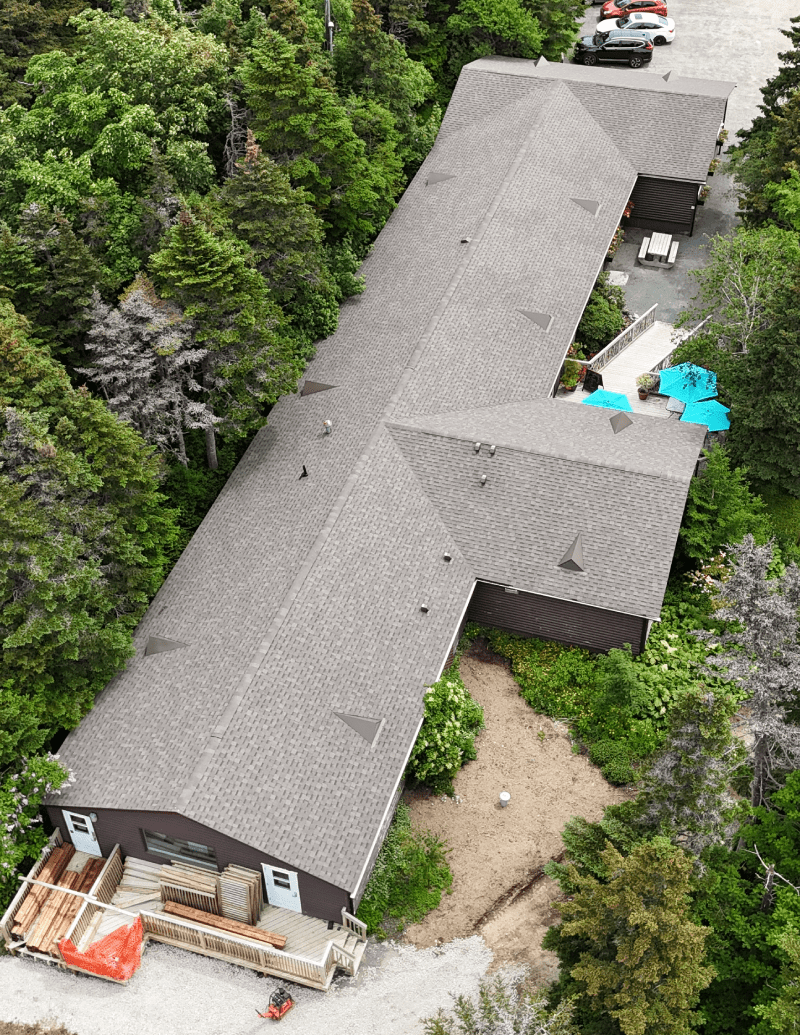 Aerial view of a T-shaped building with a shingled roof surrounded by trees, featuring a pool area Aerial view of a T-shaped building with a shingled roof surrounded by trees, featuring a pool area